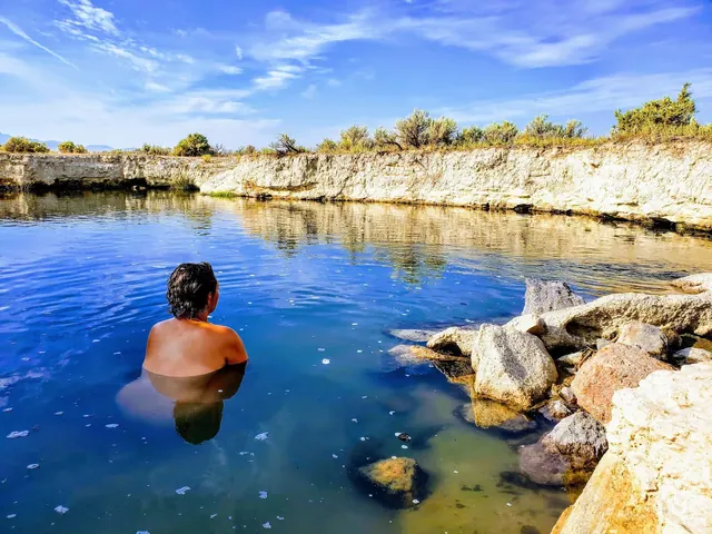 Person relaxing in Bog Hot Springs, an outdoor natural pool near Denio, Nevada, under a clear blue sky with rocky edges and shrubs