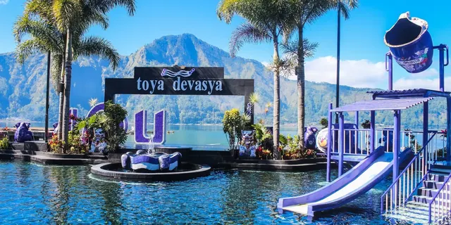 Outdoor mineral pool at Toya Devasya surrounded by palm trees and a sign with mountains visible in the background on a sunny day.