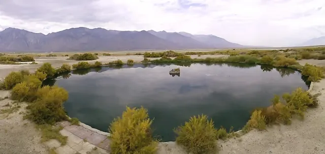 The calm water of Dirty Sock Hot Springs reflects cloudy skies and yellow-green shrubs with distant mountain ranges on the horizon close to.