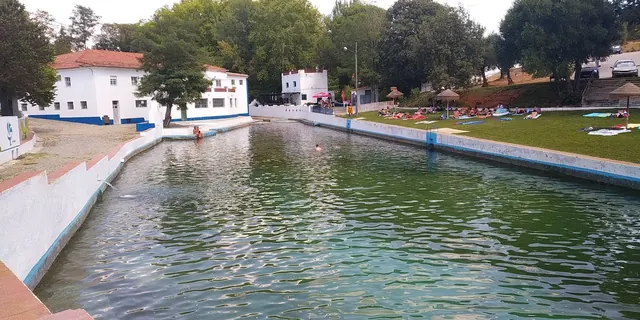 A long, rectangular outdoor hot spring pool with clear green water, surrounded by trees and white buildings at Termas da Sulfurea in Fronte.