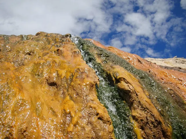Water cascades down orange and green mineral formations under a partly cloudy sky at Buckeye Hot Springs.