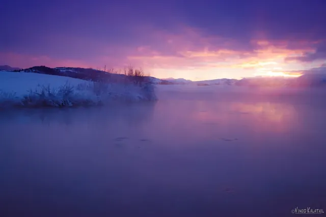Steam rises from Kelly Hot Springs water at sunset with snow-covered bushes under a purple and orange sky in Wyoming.