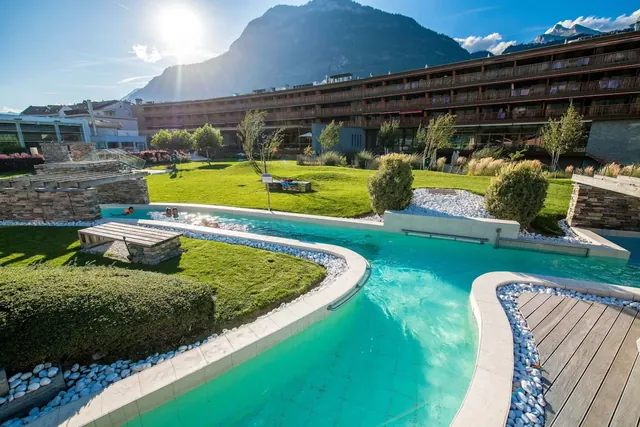 Sunlit mineral water pool with wooden benches and stone walls at Bains de Saillon near Martigny surrounded by grass and alpine peaks.