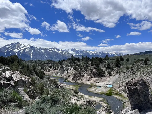 Wide view of Hot Creek Geologic Site valley with snow capped mountains in the distance and scattered trees on hillsides.