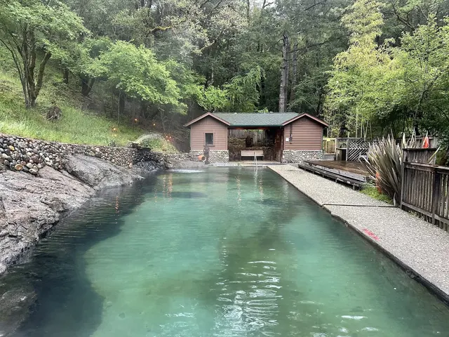 Clear water in the natural mineral pool of Orr Hot Springs reflects green trees and a rustic shelter in Ukiah, California.