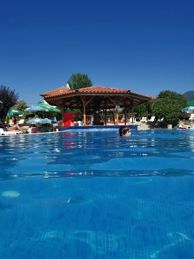 Outdoor rectangular pool with swimmers and sunbathers on white lounge chairs under a shaded pergola in Velingrad.