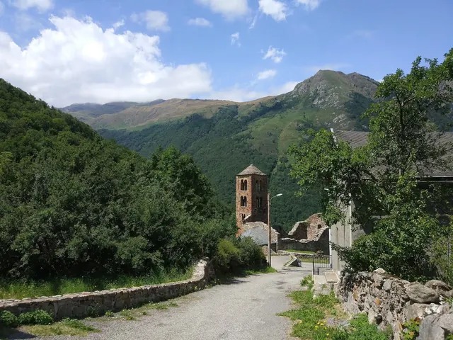 Narrow paved road with stone walls leading to an old brick tower surrounded by dense greenery and mountains in Merens-les-Vals, France.