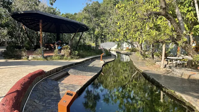 Narrow hot spring water channel bordered by stone paths and shaded seating areas under trees at Kantang Hot Spring Forest Park, Thailand.