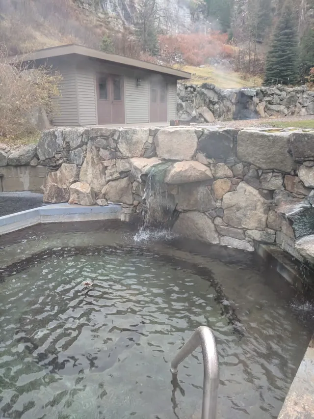 Small round soaking pool with stone wall waterfall feature and metal handrail steaming at Sleeping Child Hot Springs montana.