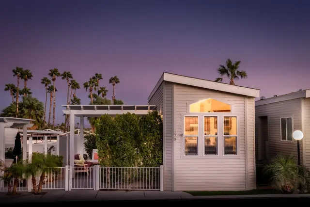 Exterior of a gray single-story cabin with lit windows and a small gated porch surrounded by palm trees at Caliente Springs Resort.