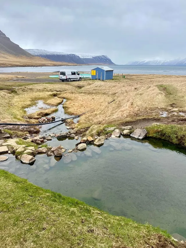 Natural hot spring pool next to small buildings, with ocean and mountain views in the background near the Reykjafjardarlaug Hot Pool in Ice.