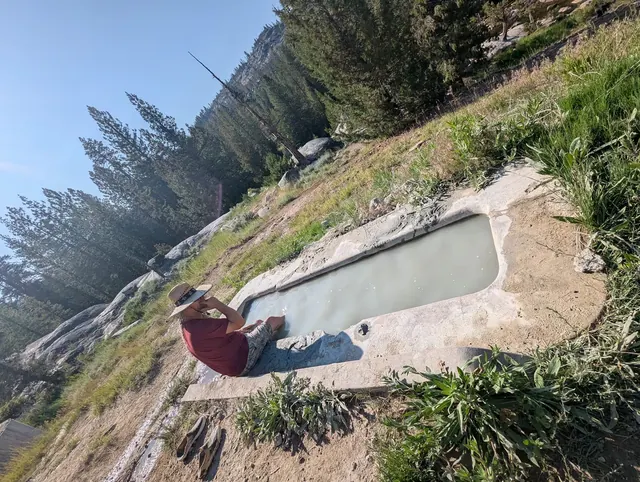 Person wearing a hat sits at the edge of a long rectangular mineral pool at Mono Hot Springs with pine trees and a rocky hillside nearby.
