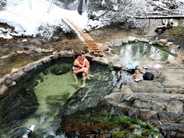 Couple embracing in the warm stone pools of Trail Creek Hot Springs near Cascade, Idaho, surrounded by light snow and a narrow wood bridge.