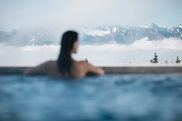 Woman relaxing in an outdoor hot spring pool with a misty mountain view at Mineralbad & Spa Rigi Kaltbad in Switzerland.