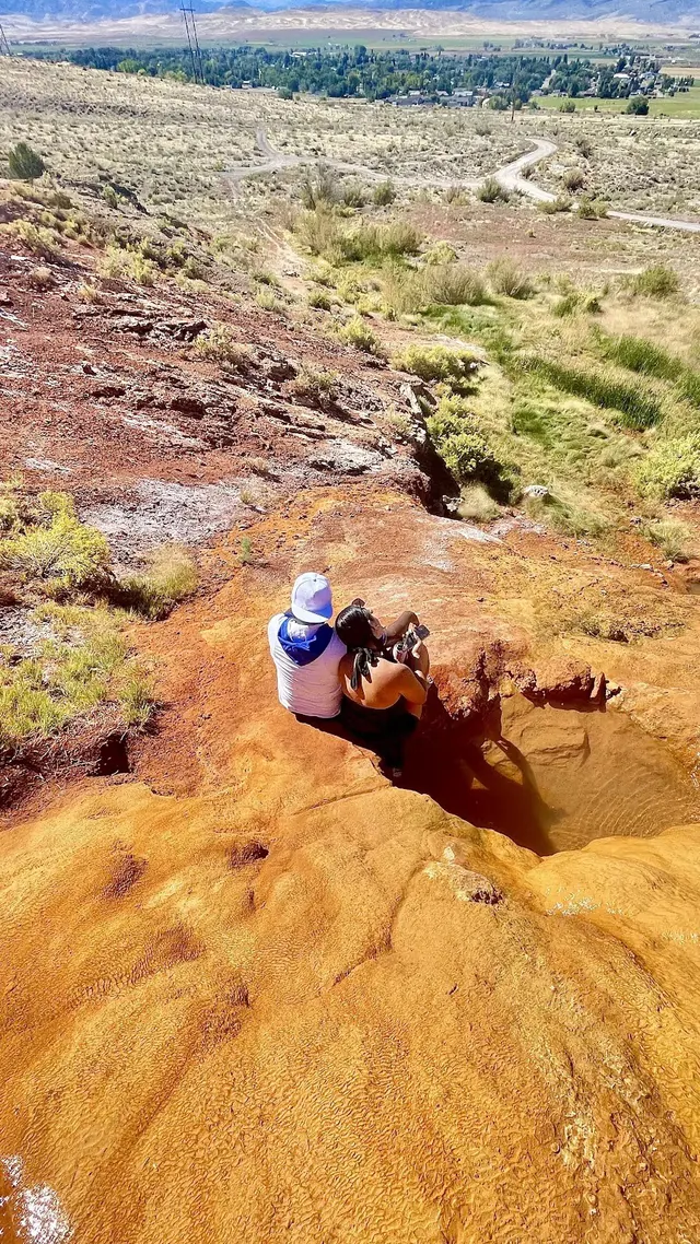 Two people sitting beside a small mineral pool on an orange rocky hillside with a view of Monroe town and distant mountains in Utah.