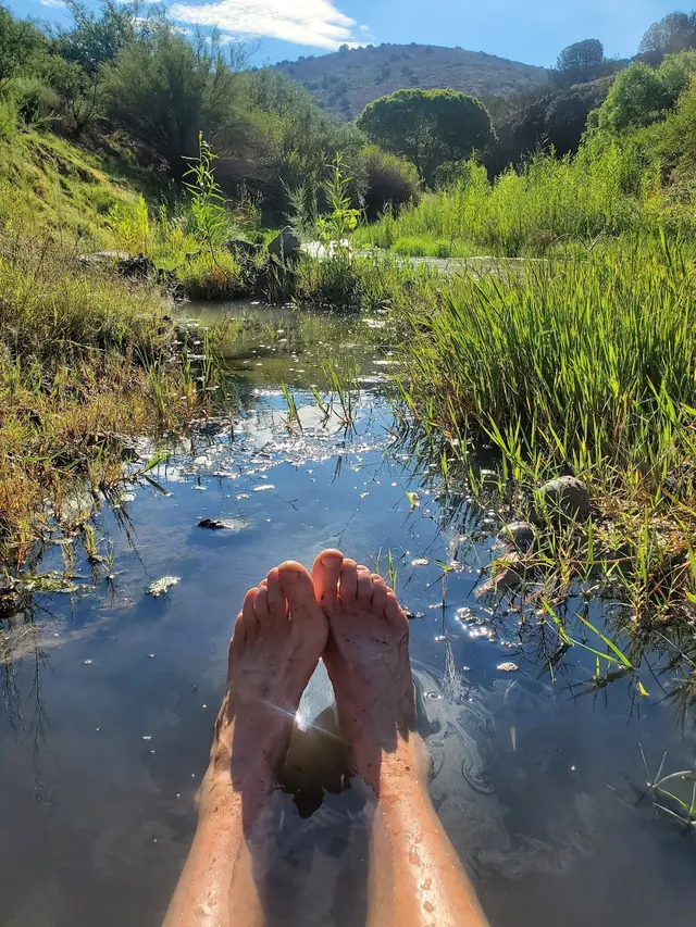 Legs and feet soaking in a shallow stream with green vegetation, hills, and a bright sky visible at San Francisco Hot Springs near Reserve.