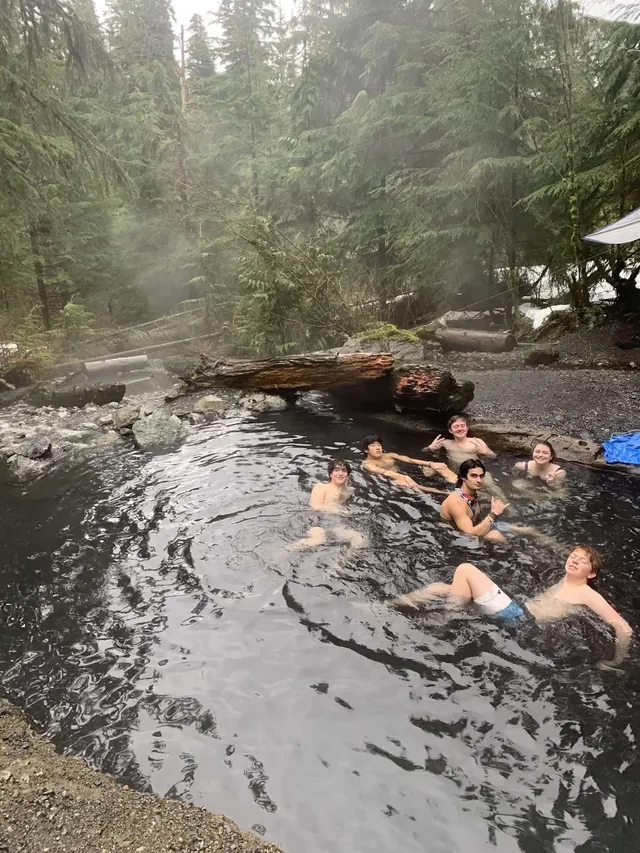 Six people relaxing in a steamy outdoor hot spring pool surrounded by dense evergreen trees at Baker Hot Springs near Glacier, Washington.