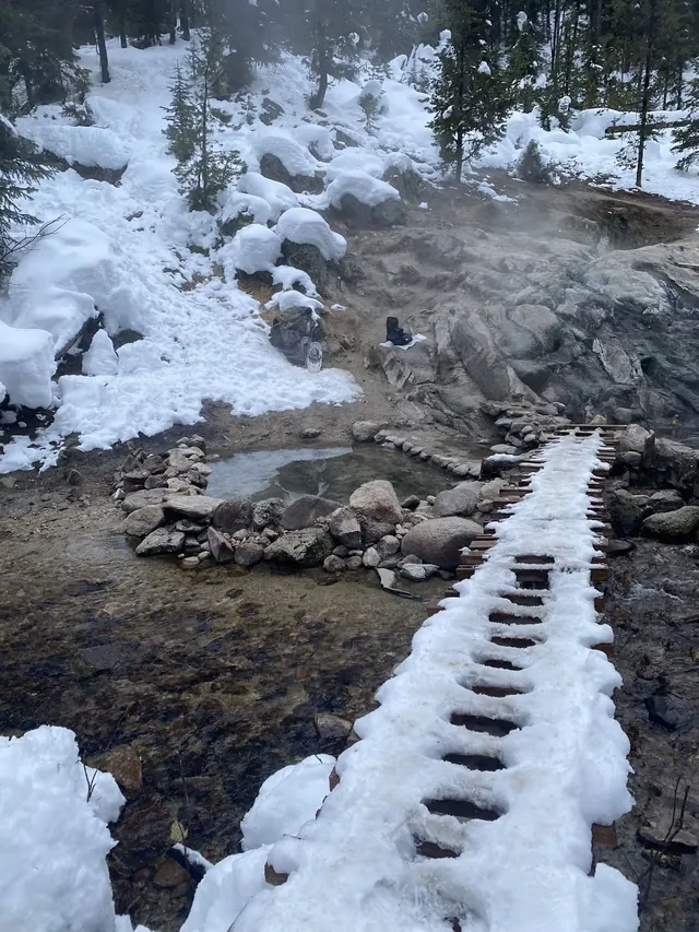 Snow-covered wooden boardwalk leading to steaming stone pools at Trail Creek Hot Springs amid snowy forest near Cascade, Idaho.