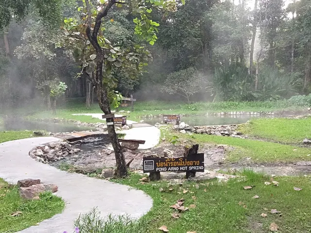 Natural hot spring area with steam rising over water pools and multiple directional signs at Pong Arng Hot Springs, Thailand.