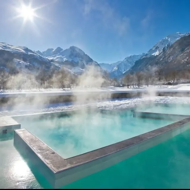 Outdoor rectangular hot spring pool at Balnea with steam rising and snow-covered mountains near Loudenvielle, France in bright sunlight.