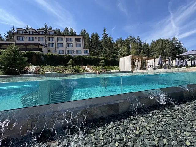 Outdoor transparent-walled hot spring pool at Pulse Therme Banya showing water cascading over stones with trees and a white building nearby.