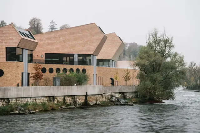 Exterior view of FORTYSEVEN Wellness-Therme showing its angular stone building along a riverside with trees nearby in Baden, Switzerland.