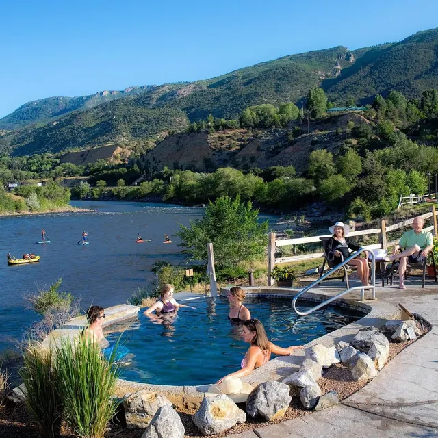 People relaxing in an outdoor hot spring pool by a river at Iron Mountain Hot Springs near Glenwood Springs, Colorado on a sunny day
