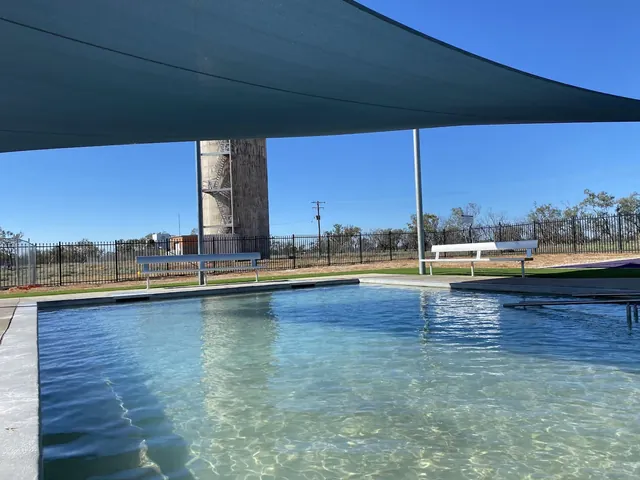 Clear water fills the outdoor soaking pool at Great Artesian Bore Baths with benches, shade sails, and a tall water tower visible nearby in.