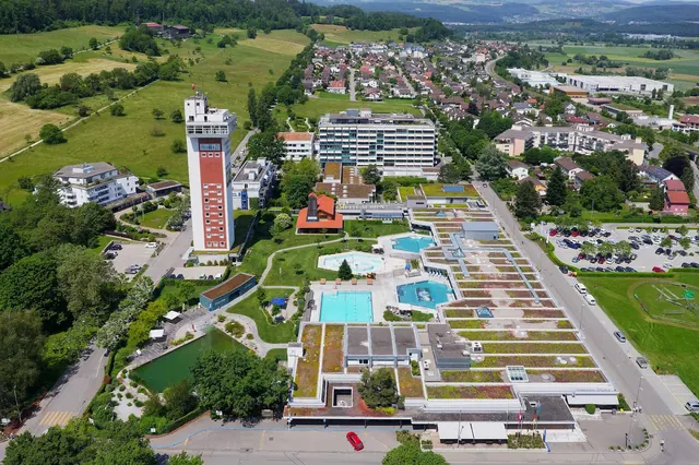 Wide aerial perspective of Therme Zurzach complex showing multiple pools, green roof sections, nearby residential area, and hills.