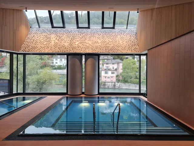 Indoor soaking pool with large windows overlooking a riverside view and modern wooden ceiling design at FORTYSEVEN Wellness-Therme in Baden.