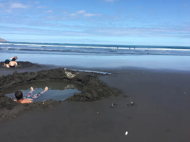 Man soaking in a natural hot spring pool dug in black sand near the ocean shore at Kawhia Hot Springs in Kawhia New Zealand.