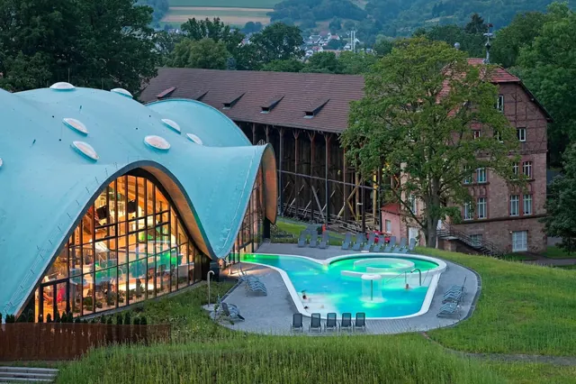 Daytime scene of outdoor mineral pool at Toskana Therme Bad Orb near large curved glass building and green landscape.