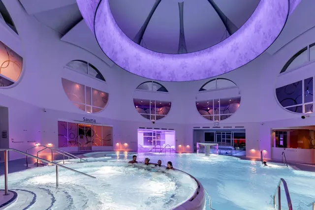 Interior view of an indoor hot spring pool illuminated with purple ambient lights under a large dome ceiling at Bogn Engiadina Scuol.