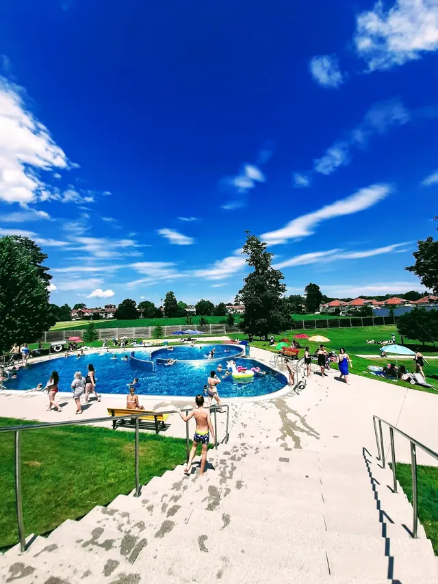 Outdoor public pool facility with people swimming and walking on stairs under blue sky near trees and houses in Bad Aibling, Germany.