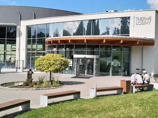 Modern entrance building of Therme Losiny in Velke Losiny Czechia with glass facade reflecting sky and a planted area nearby.