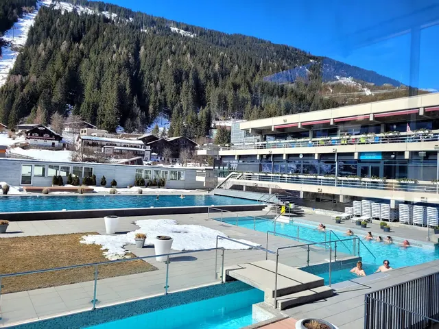 Outdoor and indoor pools at Felsentherme Bad Gastein featuring swimmers, snow patches, modern buildings, and pine-covered mountain views.
