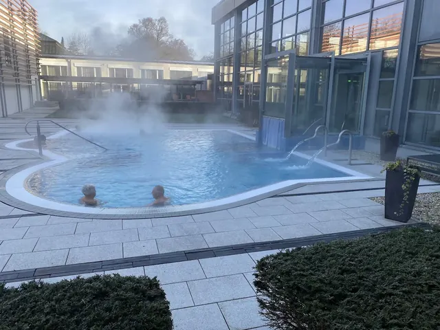Steam rises from a modern outdoor hot spring pool at Therme Bad Steben with bathers enjoying the warm waters under a crisp sky.