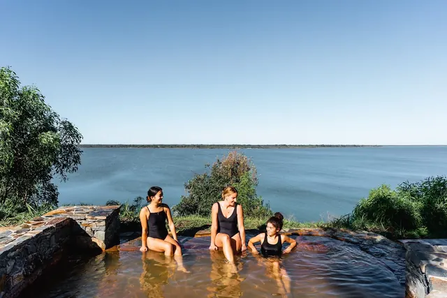 Three women sitting in a large stone soaking pool overlooking a wide body of water at Metung Hot Springs near Bairnsdale, Australia.