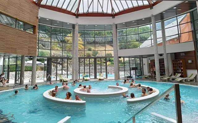 Indoor circular hot spring pool at Les Bains du Rocher in Cauterets with large windows showing outdoor area and many visitors.