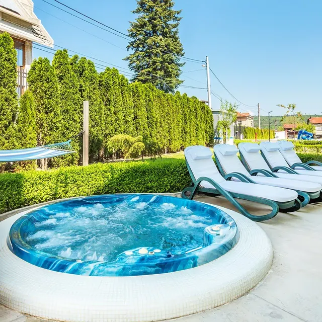 Blue bubbling hot tub with several white cushioned lounge chairs lined up next to it on a concrete patio under a clear sky near Dolna Banya.
