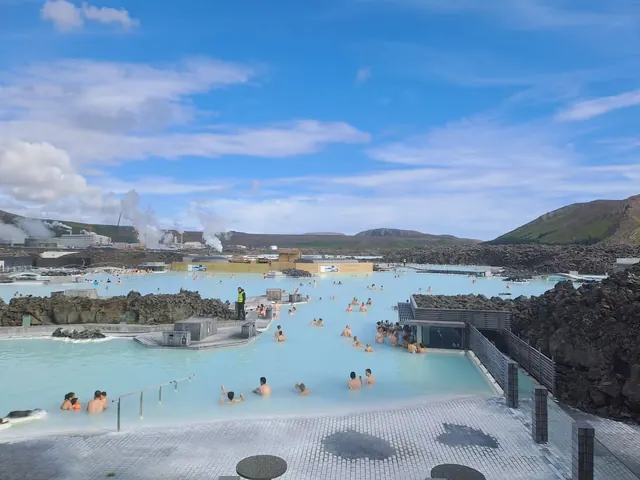 Blue Lagoon hot spring pool with milky blue water and people relaxing near black lava rocks under a clear blue sky in Iceland.