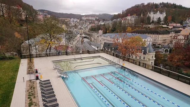 Two outdoor soaking pools with a fenced deck area and black lounge chairs overlooking the town of Karlovy Vary in Czechia during fall.