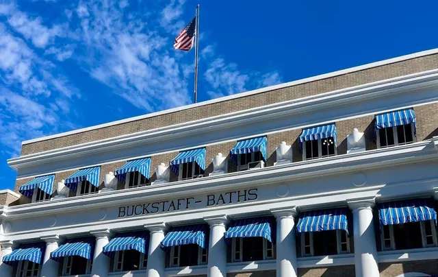 Buckstaff Bathhouse in Hot Springs Arkansas features white columns and blue striped awnings under a partly cloudy sky with an American flag.