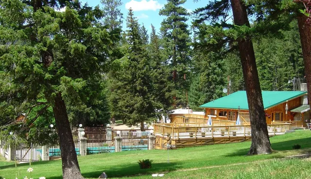 Outdoor spa area with multiple wooden buildings and tall pine trees near a fenced pool at Lost Trail Hot Springs in Montana.