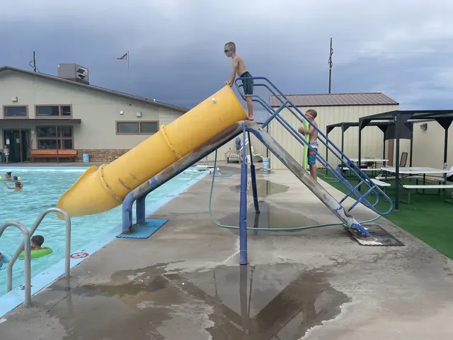 Children playing by a yellow water slide beside the outdoor pool at Splashland Hot Springs near Alamosa, Colorado, under a cloudy sky.
