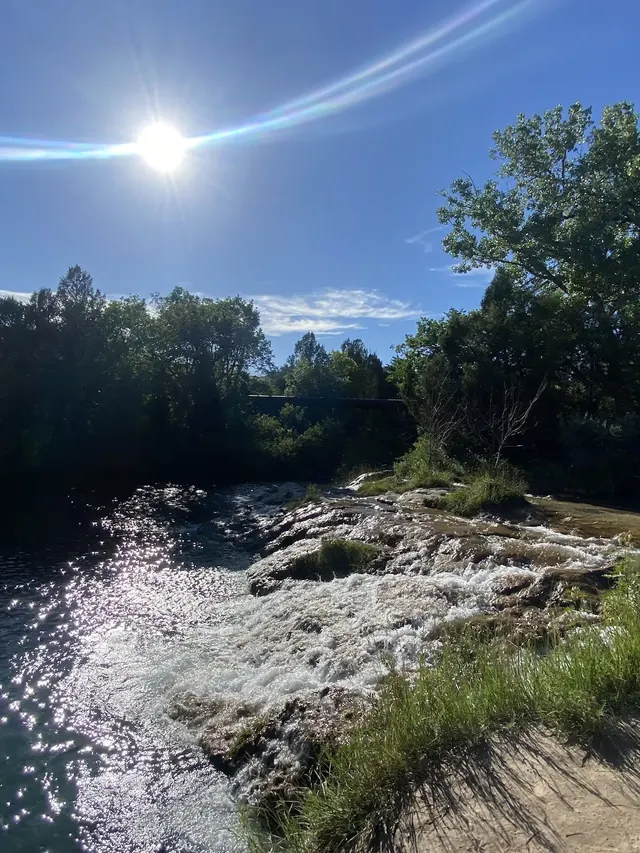 Sunlight reflects off rippling water flowing over smooth rocks with surrounding trees casting shadows at Cascade Falls in Hot Springs south.