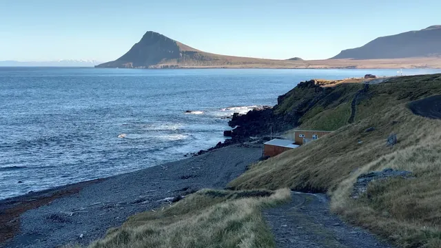 Coastal hillside view down to Krossnes Swimming Pool with ocean waters and a large mountain in the distance near Nordurfjordur, Iceland.
