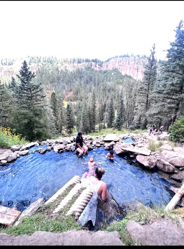 Group of people sit and soak in a rock-bordered hot spring pool high on a hillside with dense forest and red rock cliffs near Jemez Springs.