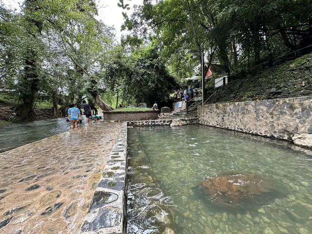 People walking beside clear water pools at Hin Dad Hot Spring with lush green trees in Thong Pha Phum, Nan, Thailand.