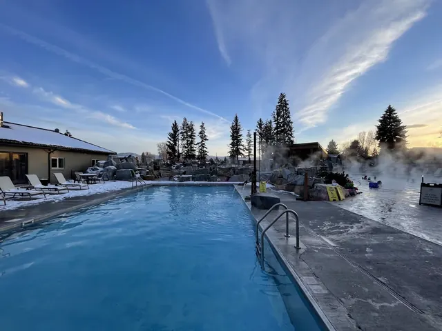 Steaming outdoor pool at Bozeman Hot Springs with snow on the ground and pine trees visible under a cloudy sky.